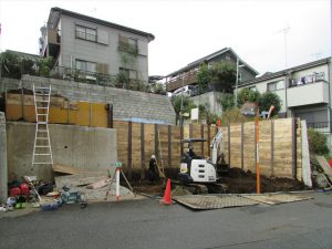 3.5ｍの擁壁の一部を解体して掘削を先行しながら平地を作り地盤補強杭を貫入します。さらに掘り進んで整地を行い重機を入れて山留杭打設してさらに掘削して道路レベル-50cmの杭頭を露出して掘削を完了します