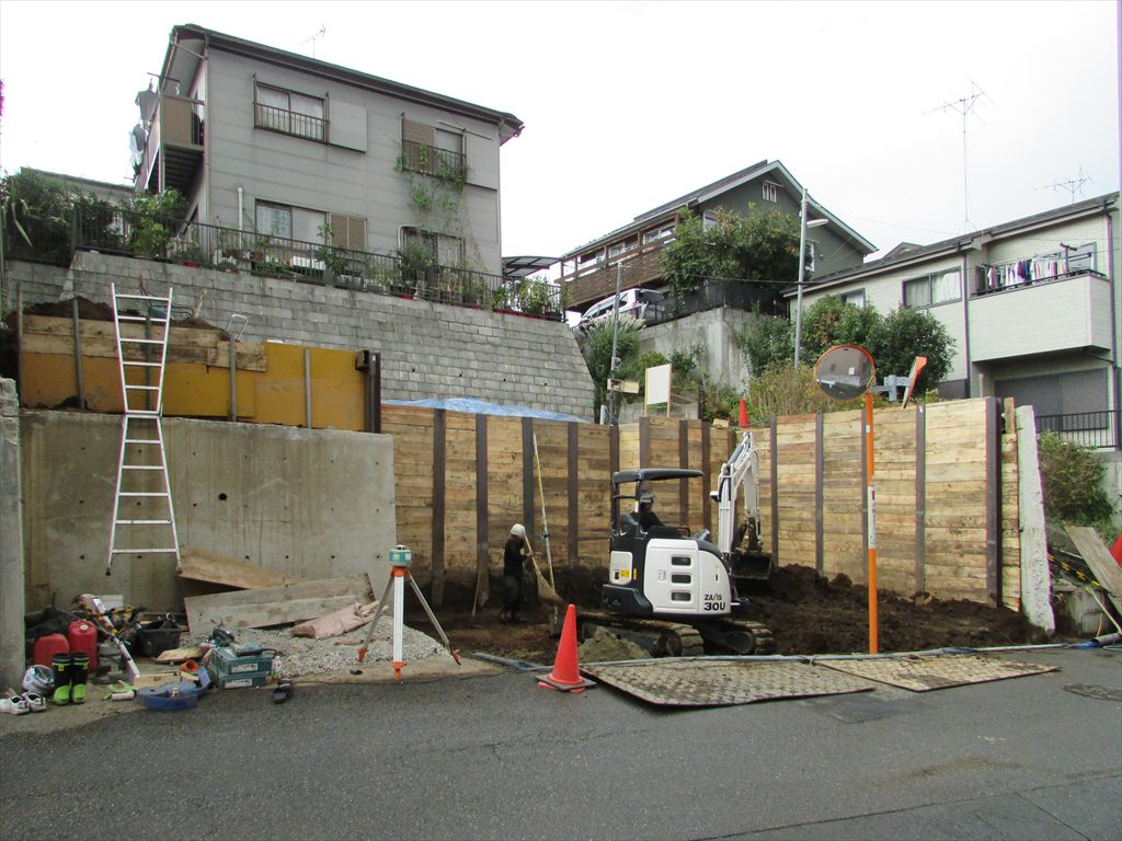 3.5ｍの擁壁の一部を解体して掘削を先行しながら平地を作り地盤補強杭を貫入します。さらに掘り進んで整地を行い重機を入れて山留杭打設してさらに掘削して道路レベル-50cmの杭頭を露出して掘削を完了します